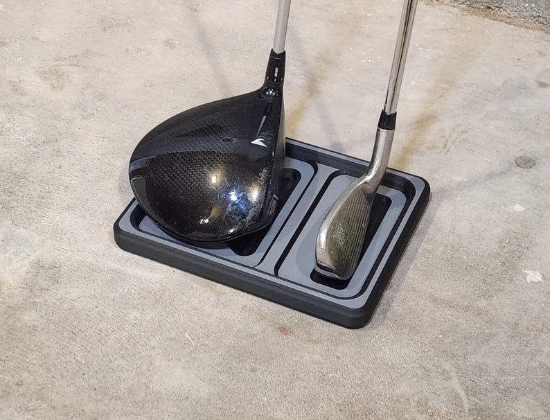 May include: A black and gray golf club organizer holding a black driver and a silver iron. The organizer has two compartments and is sitting on a concrete surface.