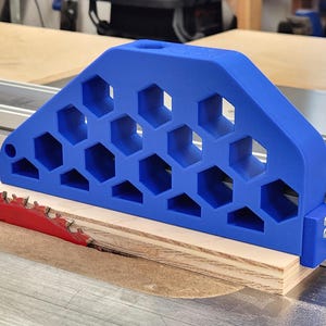 Puede incluir: Una guía de corte azul impresa en 3D con un patrón de panal, colocada contra una guía metálica en una sierra de mesa. La herramienta presenta una serie de recortes hexagonales y está situada junto a una hoja de sierra roja cortando madera clara.