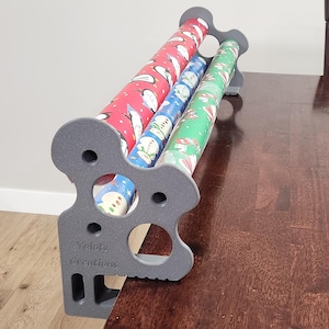 May include: A gray 3D-printed wrapping paper organizer with the text "Yelotz Creations" embossed on the side. The organizer holds three rolls of holiday-themed wrapping paper in red, blue, and green, and is designed to clamp onto a table edge.