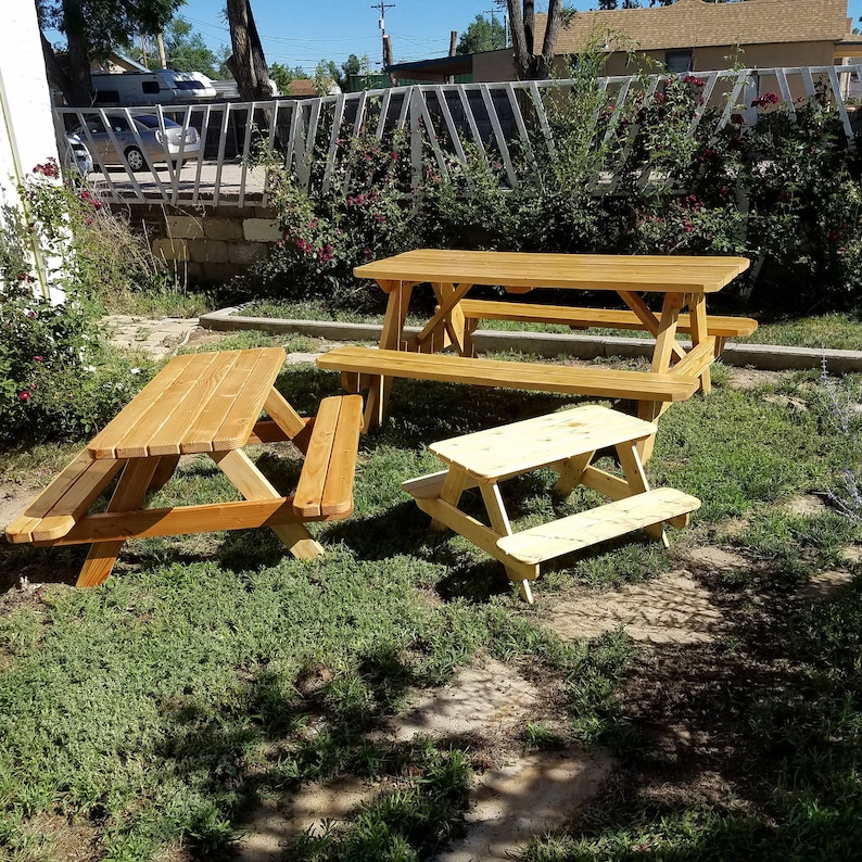 May include: Three wooden picnic tables in a grassy backyard. The largest table is in the center, with two smaller tables to the left and right. All tables are made of natural wood and have a rustic look.