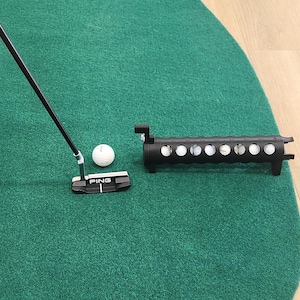 Könnte beinhalten: Ein schwarzer Ping-Putter mit weißer Ausrichtungslinie, ein einzelner weißer Golfball und ein schwarzes zylindrisches Golfball-Aufbewahrungsrohr mit mehreren kreisförmigen Aussparungen, die mehrere weiße Golfbälle enthalten, auf einer grünen Fläche.