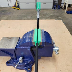 May include: A blue metal bench vise holding a black golf club shaft with a green plastic alignment tool attached to the shaft. The vise features a silver handle and a flat anvil surface, while the green tool is positioned at the top of the shaft.