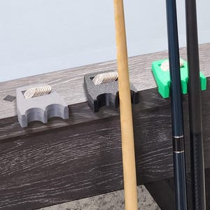 May include: Three 3D-printed pool cue holders in gray, black, and green, each holding a stack of coins. A wooden pool cue and two other cues are also visible. The holders are placed on the edge of a dark wood pool table.