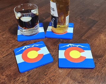 May include: Four square coasters with the Colorado state flag design. Each coaster features a blue background with a white stripe, a red and yellow emblem, and a mountain range. A glass of dark liquid and a beer bottle rest on two of the coasters.