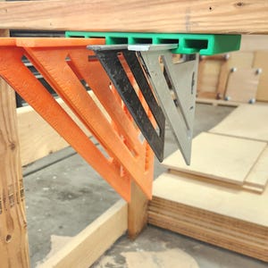 Puede incluir: Un estante de almacenamiento de plástico verde montado en la parte inferior de un banco de trabajo de madera, que sostiene dos escuadras de plástico naranja, una escuadra de metal negro y una escuadra de metal plateado, colgando verticalmente.