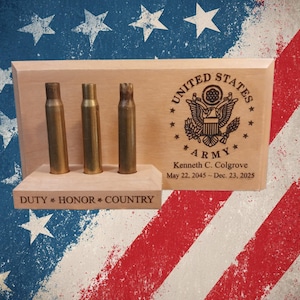 May include: A light-colored wooden display stand featuring three brass bullet casings. The stand is engraved with the United States Army emblem, the name Kenneth C. Colgrove, the dates May 22, 2045 - Dec. 23, 2025, and the text DUTY * HONOR * COUNTRY.