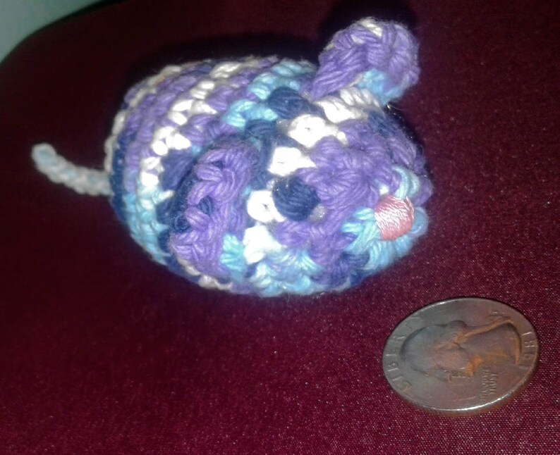 May include: A crocheted purple, blue, and white cat toy shaped like a mouse. The toy has a pink nose and a white tail.