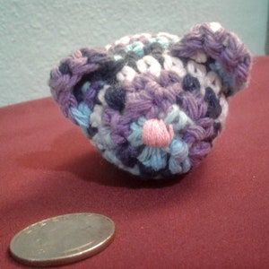 May include: A small crocheted stuffed animal in the shape of a bear. The bear is made with purple, blue, white, and pink yarn. The bear has a pink nose and is sitting on a red surface.