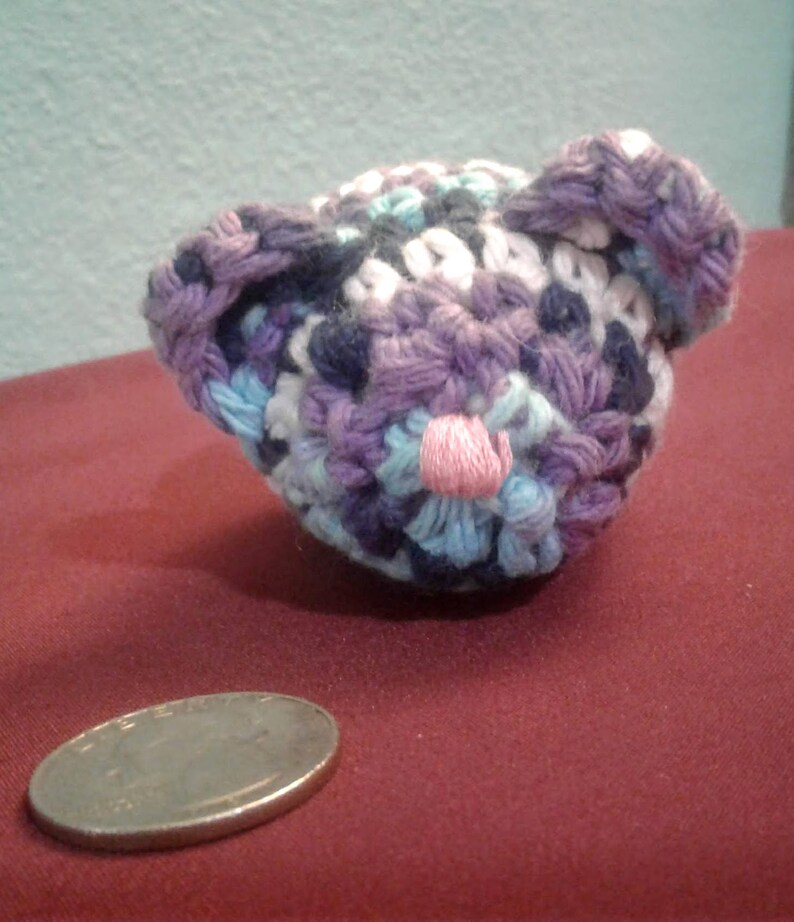 May include: A small crocheted stuffed animal in the shape of a bear. The bear is made with purple, blue, white, and pink yarn. The bear has a pink nose and is sitting on a red surface.