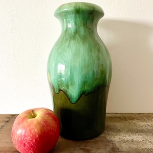 May include: A tall, green ceramic vase with a glossy finish. The vase has a wide base that tapers to a narrower neck and flared rim. A red apple sits to the left of the vase. The vase is approximately 30 cm tall.