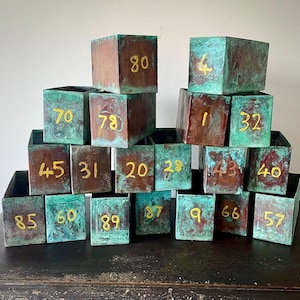May include: A display of copper boxes with a green verdigris finish. Each box is marked with a gold number, from 1 to 89. The boxes are arranged on a dark surface, showcasing their aged appearance.