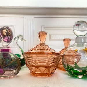 Könnte beinhalten: Sammlung von Vintage-Parfümflaschen aus Glas und einer Schale mit Deckel. Die Flaschen haben dekorative Verschlüsse und bemalte Designs in Lila, Grün und Orange. Die Schale mit Deckel ist pfirsichfarben.
