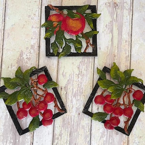 Könnte beinhalten: Drei dekorative Wandkunstwerke mit Früchten. Ein quadratischer Rahmen hält rote Äpfel mit grünen Blättern, während zwei rautenförmige Rahmen Trauben roter Kirschen mit grünen Blättern zeigen. Auf einem verwitterten Holzhintergrund.