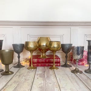 May include: A collection of antique goblets and chalices in various metallic finishes, including silver, gold, and pewter. The goblets are displayed on a wooden surface, with some resting on red books. The collection includes ornate and simple designs.