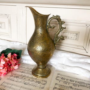 May include: A vintage brass pitcher with an ornate floral pattern and a dragon-shaped handle. The pitcher is displayed on a table with sheet music and a pink flower arrangement. The pitcher is approximately 12 inches tall.