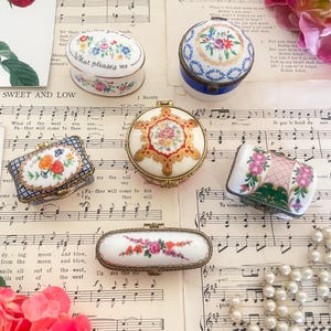 May include: An assortment of vintage porcelain trinket boxes with floral designs. The boxes vary in shape and size, featuring intricate hand-painted details in pastel colors. They rest on a backdrop of sheet music and decorative elements.