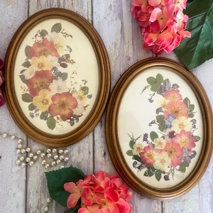 May include: Two vintage oval framed floral prints. The frames are a warm gold color. The prints feature a variety of colorful flowers in shades of red, yellow, and white. The background is a cream color.