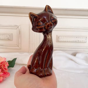 May include: A brown ceramic cat figurine with a glazed finish. The cat is sitting with its head tilted and its eyes closed.