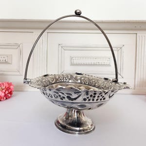 May include: Silver-plated serving basket with a handle. The basket has an ornate, openwork design with a scalloped edge and a pedestal base. The handle is arched and topped with a small ball.