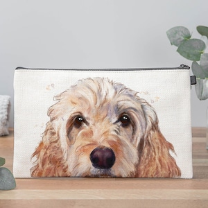 May include: A white canvas makeup bag featuring a watercolour illustration of a golden doodle dog's face. The dog has brown eyes and a pink nose.
