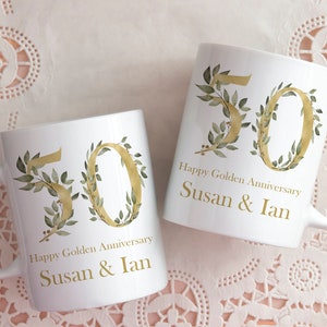May include: Two white ceramic mugs with gold lettering and green foliage designs. The mugs say "Happy Golden Anniversary Susan & Ian" and feature the number "50" in gold.