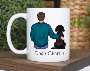 Può includere: Tazza in ceramica bianca con l'illustrazione di una persona e un cane nero. La persona indossa un maglione verde acqua e jeans blu. Il testo "Dad & Charlie" è stampato sulla tazza.