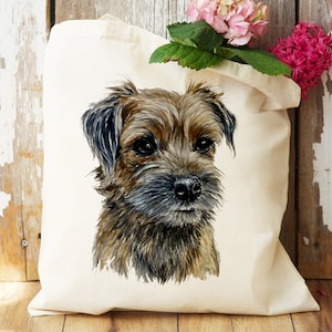 Pu&ograve; includere: Una borsa di tela bianca con un'illustrazione ad acquerello di un Border Terrier marrone e nero. Il cane guarda lo spettatore con un'espressione seria.