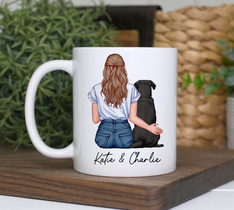 May include: White ceramic mug featuring an illustration of a woman with long brown hair and a black dog. The woman is wearing a white t-shirt and blue jeans. The text "Katie & Charlie" is printed on the mug.