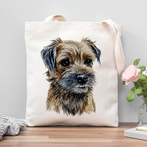 Pu&ograve; includere: Una borsa tote in tela bianca con un'illustrazione ad acquerello di un Border Terrier marrone e nero. Il cane guarda direttamente lo spettatore.