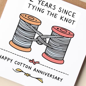 May include: A square greeting card featuring a cartoon illustration of two wooden spools of light blue thread tied together. The text reads 2 YEARS SINCE TYING THE KNOT and HAPPY COTTON ANNIVERSARY. The card is white with black line art and text.