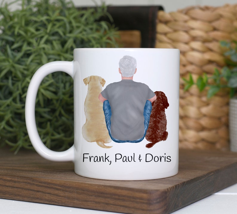 K&ouml;nnte beinhalten: Wei&szlig;e Keramiktasse mit einem Design einer Person und zwei Hunden, einem gelben Labrador und einem braunen Hund. Auf der Tasse stehen die Namen "Frank, Paul & Doris". Die Tasse hat einen wei&szlig;en Henkel.