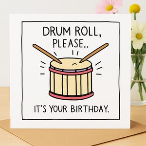 May include: A birthday card featuring a cartoon drum illustration. The drum is beige with red and black accents, and drumsticks are poised above it. The text reads "DRUM ROLL, PLEASE... IT'S YOUR BIRTHDAY."