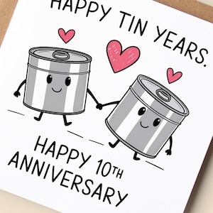 May include: A greeting card with the text "HAPPY TIN YEARS" and "HAPPY 10TH ANNIVERSARY". The card features two cartoon tin cans holding hands, with hearts above them. The card is on a brown background.