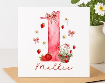 Può includere: Un biglietto d'auguri quadrato con un grande numero uno ad acquerello rosa, decorato con fragole rosse, margherite bianche e fiocchi a quadretti rossi. Un cestino pieno di fiori e fragole si trova alla base. Il nome Millie è scritto sotto.