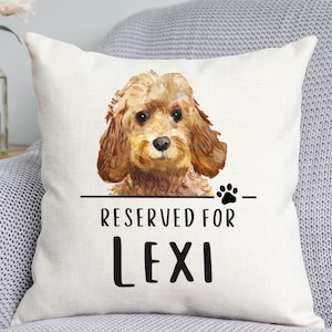 May include: A square off-white throw pillow featuring a watercolour illustration of a brown curly-haired dog. Below the dog is a horizontal line with a black paw print and the black text that reads RESERVED FOR LEXI in a bold font.