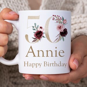 May include: White ceramic mug with a gold 50 and floral design. The text "Annie Happy Birthday" is printed on the mug.