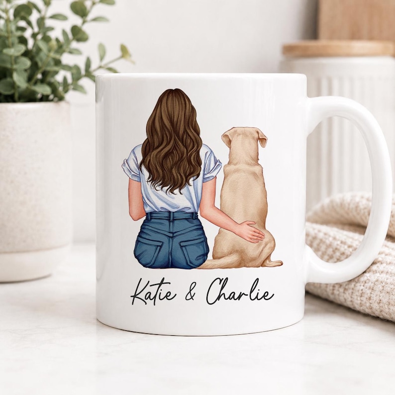 May include: A white ceramic mug featuring a printed illustration of a person with long brown hair wearing a white t-shirt and blue jeans, sitting next to a light brown dog. The text Katie & Charlie is printed in black script below the figures.