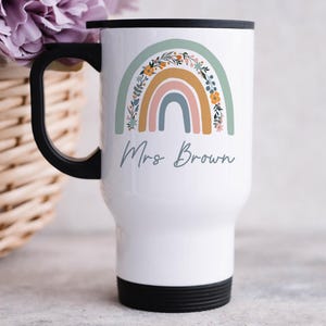 May include: A white travel mug with a black handle and lid, featuring a printed graphic of a multi-colored rainbow with floral accents in sage green, mustard yellow, and dusty pink. The text Mrs Brown is printed in a dark grey script font below.
