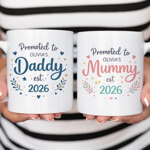 May include: Two white ceramic mugs with handles, each featuring text. One mug says "Promoted to Daddy" in blue, the other says "Promoted to Mummy" in pink. Both mugs include the name "Olivia's" and the year "2026".