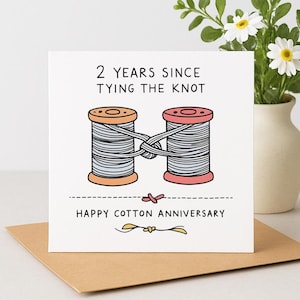 May include: A square greeting card with a white background featuring an illustration of two thread spools tied together with gray thread. The text reads 2 YEARS SINCE TYING THE KNOT and HAPPY COTTON ANNIVERSARY in black ink with a small yellow knot graphic.