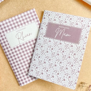 May include: Two rectangular notebooks with soft covers. One notebook features a mauve and white gingham pattern with a label reading Nodiadau Rhian. The other notebook has a mauve floral print with a label reading Nodiadau Mam.