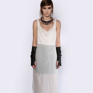 May include: A white maxi dress with a layered design. The dress has a scoop neck and a fitted bodice. The skirt is flowy and sheer. The model is wearing black leather fingerless gloves and black heeled shoes.