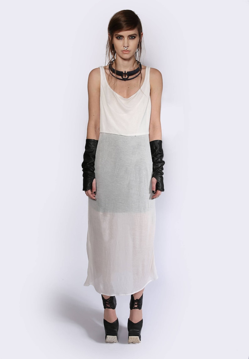 May include: A white maxi dress with a layered design. The dress has a scoop neck and a fitted bodice. The skirt is flowy and sheer. The model is wearing black leather fingerless gloves and black heeled shoes.