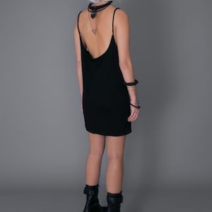 May include: A black sleeveless dress with a cowl neck and a low back. The dress is worn by a person with white hair and black platform boots.
