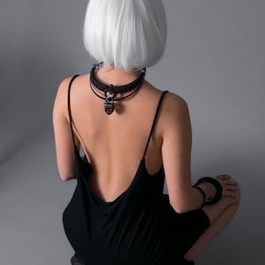 May include: A woman with short, white hair is wearing a black, sleeveless dress with a low back. She is wearing a black choker necklace with a large, brown pendant.
