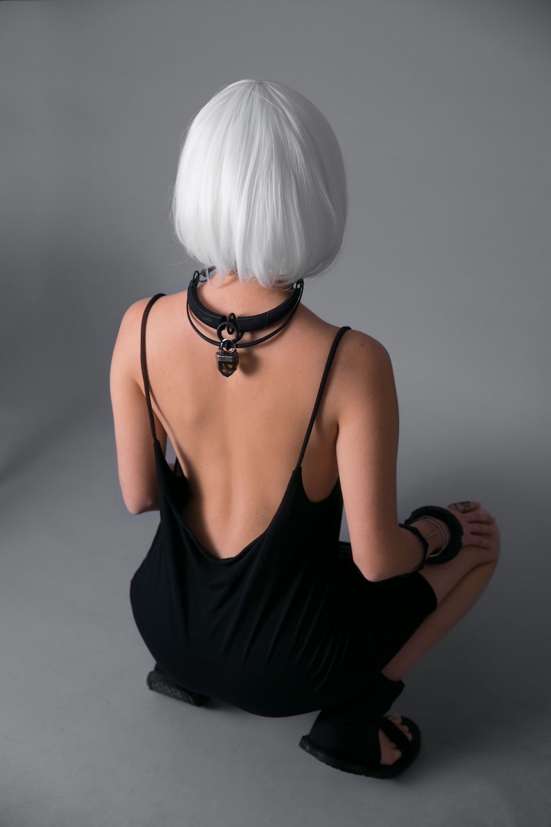 May include: A woman with short, white hair is wearing a black, sleeveless dress with a low back. She is wearing a black choker necklace with a large, brown pendant.