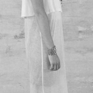 May include: A woman wearing a white sleeveless top with a sheer overlay and a silver chain necklace. She is wearing a silver bracelet on her wrist and black platform sandals.