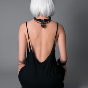 May include: A person with short, white hair wearing a black, sleeveless dress with a low back. The person is wearing a black choker with a crystal pendant.