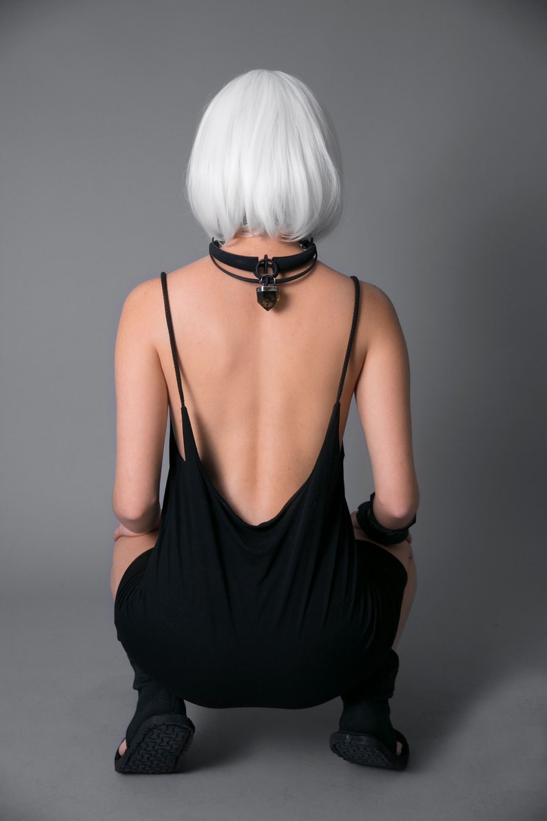 May include: A person with short, white hair wearing a black, sleeveless dress with a low back. The person is wearing a black choker with a crystal pendant.