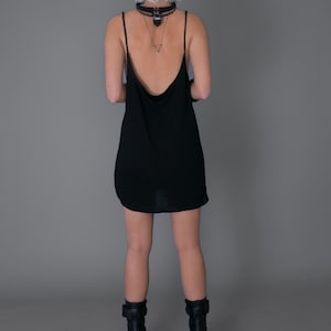 May include: A woman wearing a black sleeveless dress with a low back and black platform boots. She is wearing a silver necklace with a pendant.
